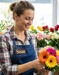 Woman in a floral shop holding a bouquet of flowers, wearing a blue apron with 'Susan' on it.
