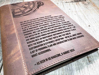 A brown, custom-engraved leather notebook cover featuring a geometric unicorn head design. The notebook is resting on an antique-style, ornate wooden desk alongside a glass inkwell and a feathered quill pen, creating a sophisticated and vintage aesthetic.