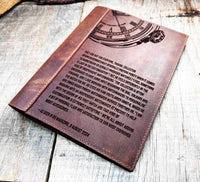 A brown, custom-engraved leather notebook cover featuring a geometric unicorn head design. The notebook is resting on an antique-style, ornate wooden desk alongside a glass inkwell and a feathered quill pen, creating a sophisticated and vintage aesthetic.