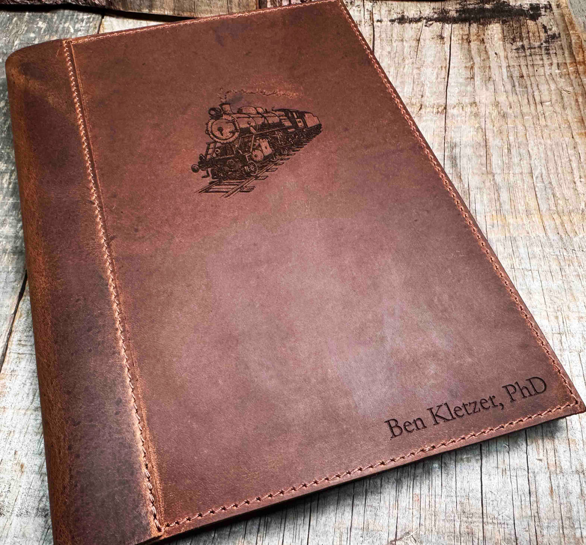 A brown, custom-engraved leather notebook cover featuring a geometric unicorn head design. The notebook is resting on an antique-style, ornate wooden desk alongside a glass inkwell and a feathered quill pen, creating a sophisticated and vintage aesthetic.