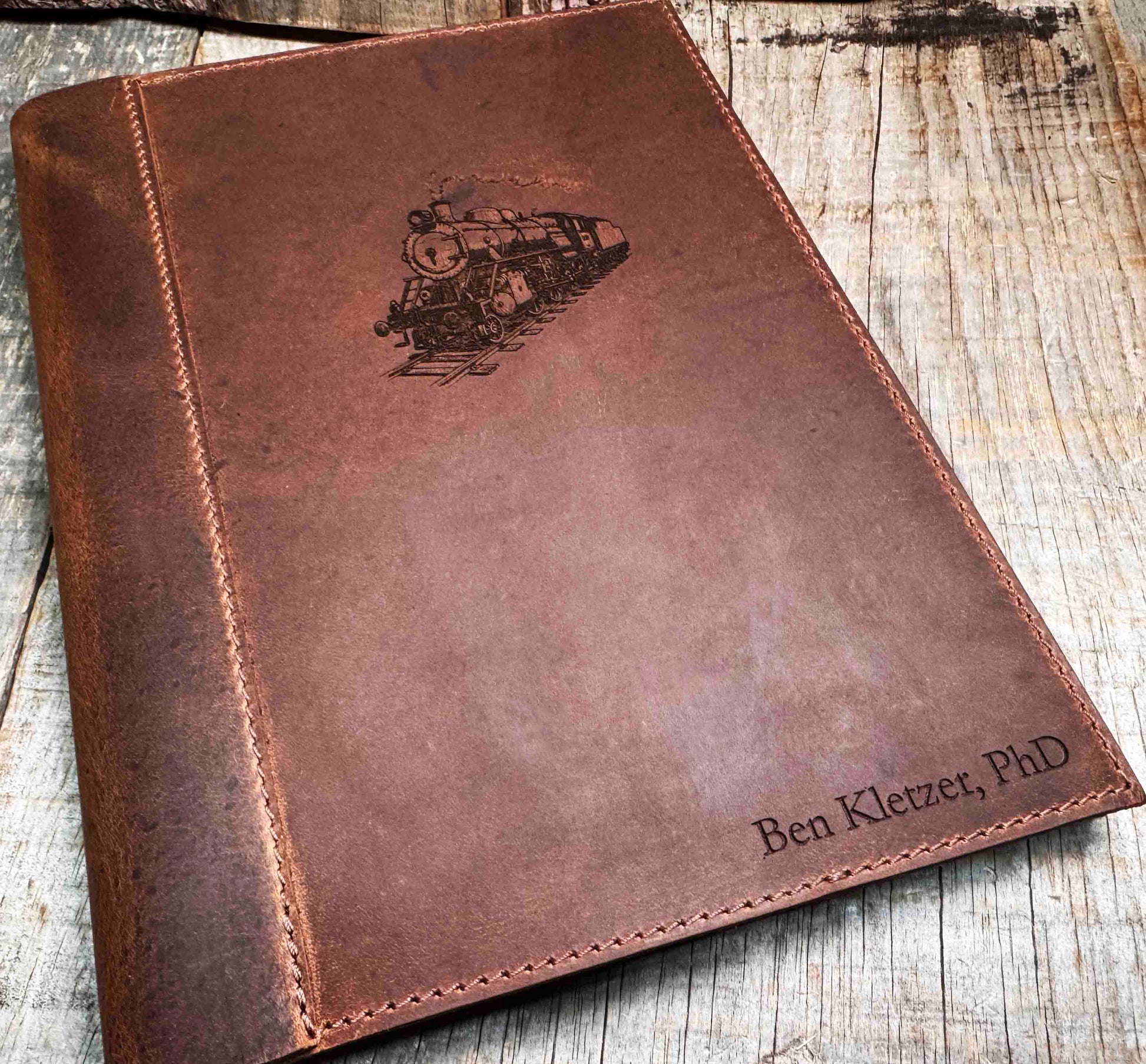 A brown, custom-engraved leather notebook cover featuring a geometric unicorn head design. The notebook is resting on an antique-style, ornate wooden desk alongside a glass inkwell and a feathered quill pen, creating a sophisticated and vintage aesthetic.