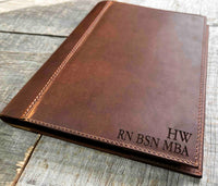 A brown, custom-engraved leather notebook cover featuring a geometric unicorn head design. The notebook is resting on an antique-style, ornate wooden desk alongside a glass inkwell and a feathered quill pen, creating a sophisticated and vintage aesthetic.