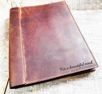 A brown, custom-engraved leather notebook cover featuring a geometric unicorn head design. The notebook is resting on an antique-style, ornate wooden desk alongside a glass inkwell and a feathered quill pen, creating a sophisticated and vintage aesthetic.