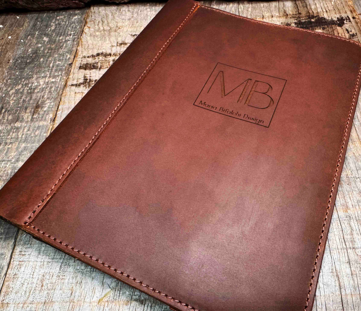 A brown, custom-engraved leather notebook cover featuring a geometric unicorn head design. The notebook is resting on an antique-style, ornate wooden desk alongside a glass inkwell and a feathered quill pen, creating a sophisticated and vintage aesthetic.