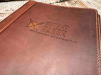 A brown, custom-engraved leather notebook cover featuring a geometric unicorn head design. The notebook is resting on an antique-style, ornate wooden desk alongside a glass inkwell and a feathered quill pen, creating a sophisticated and vintage aesthetic.