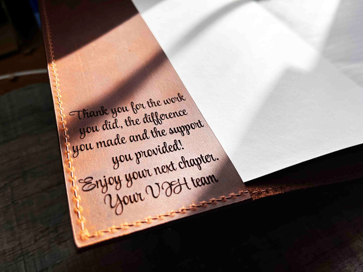 A brown, custom-engraved leather notebook cover featuring a geometric unicorn head design. The notebook is resting on an antique-style, ornate wooden desk alongside a glass inkwell and a feathered quill pen, creating a sophisticated and vintage aesthetic.