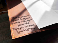 A brown, custom-engraved leather notebook cover featuring a geometric unicorn head design. The notebook is resting on an antique-style, ornate wooden desk alongside a glass inkwell and a feathered quill pen, creating a sophisticated and vintage aesthetic.