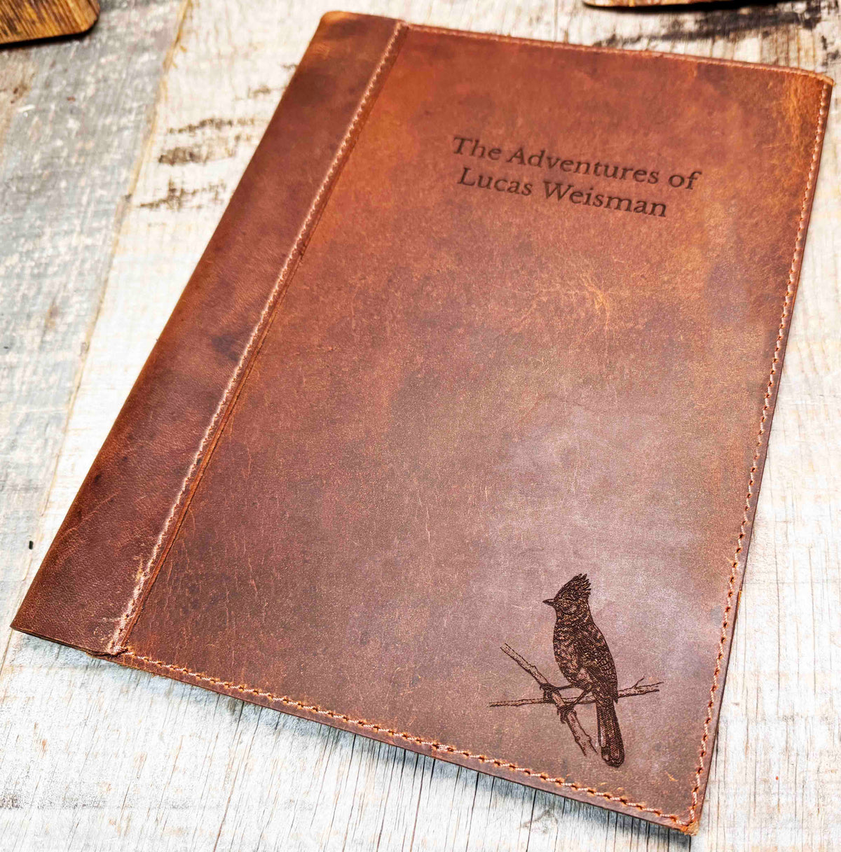 A brown, custom-engraved leather notebook cover featuring a geometric unicorn head design. The notebook is resting on an antique-style, ornate wooden desk alongside a glass inkwell and a feathered quill pen, creating a sophisticated and vintage aesthetic.