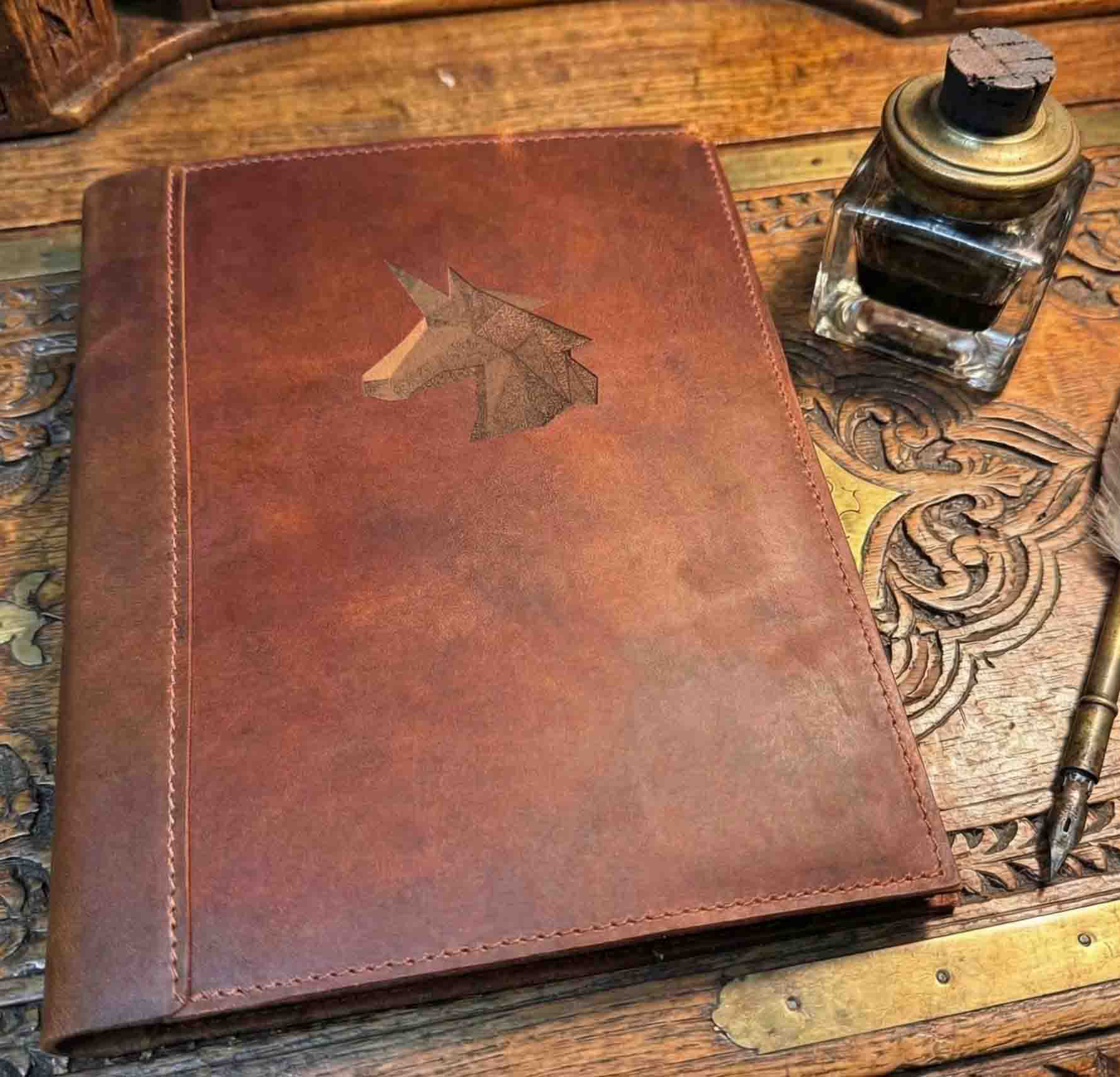 A brown, custom-engraved leather notebook cover featuring a geometric unicorn head design. The notebook is resting on an antique-style, ornate wooden desk alongside a glass inkwell and a feathered quill pen, creating a sophisticated and vintage aesthetic.