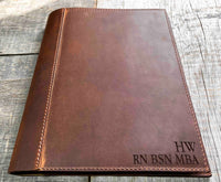 A brown, custom-engraved leather notebook cover featuring a geometric unicorn head design. The notebook is resting on an antique-style, ornate wooden desk alongside a glass inkwell and a feathered quill pen, creating a sophisticated and vintage aesthetic.