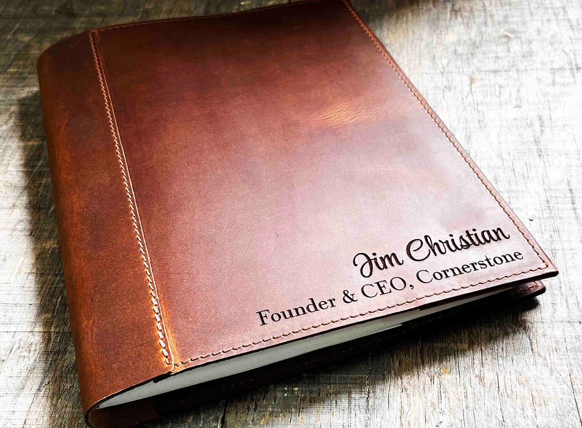 A brown, custom-engraved leather notebook cover featuring a geometric unicorn head design. The notebook is resting on an antique-style, ornate wooden desk alongside a glass inkwell and a feathered quill pen, creating a sophisticated and vintage aesthetic.