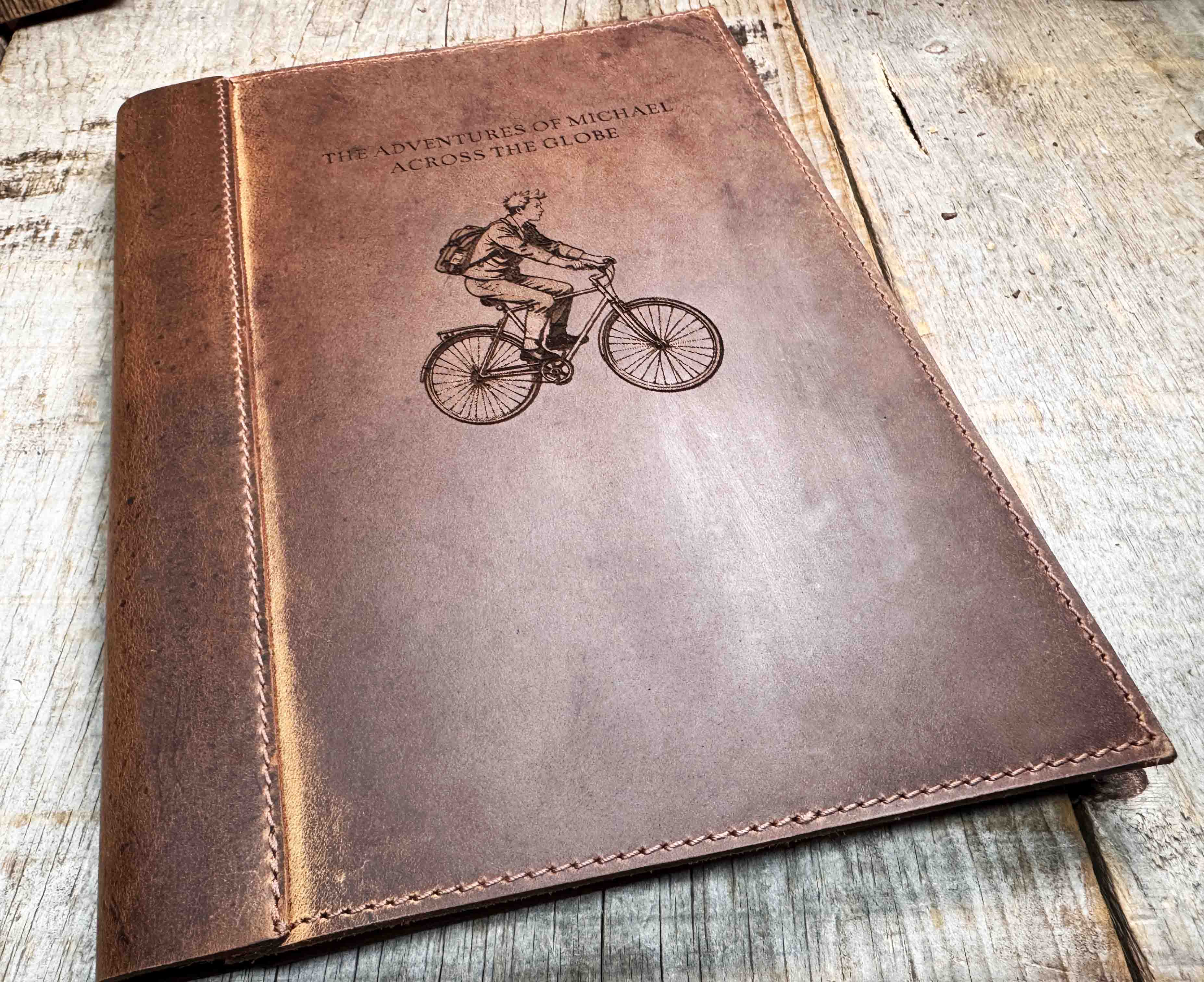 A brown, custom-engraved leather notebook cover featuring a geometric unicorn head design. The notebook is resting on an antique-style, ornate wooden desk alongside a glass inkwell and a feathered quill pen, creating a sophisticated and vintage aesthetic.