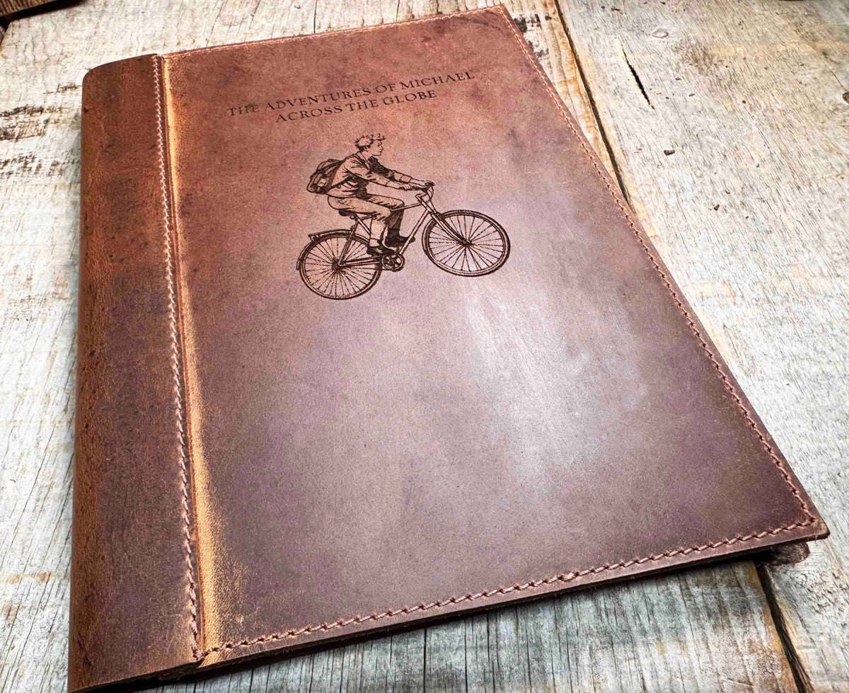 A brown, custom-engraved leather notebook cover featuring a geometric unicorn head design. The notebook is resting on an antique-style, ornate wooden desk alongside a glass inkwell and a feathered quill pen, creating a sophisticated and vintage aesthetic.