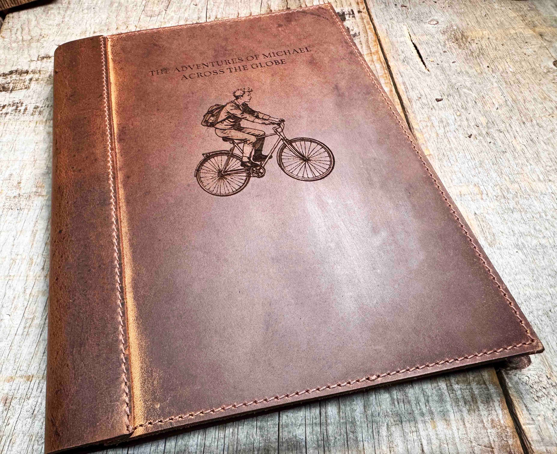 A brown, custom-engraved leather notebook cover featuring a geometric unicorn head design. The notebook is resting on an antique-style, ornate wooden desk alongside a glass inkwell and a feathered quill pen, creating a sophisticated and vintage aesthetic.