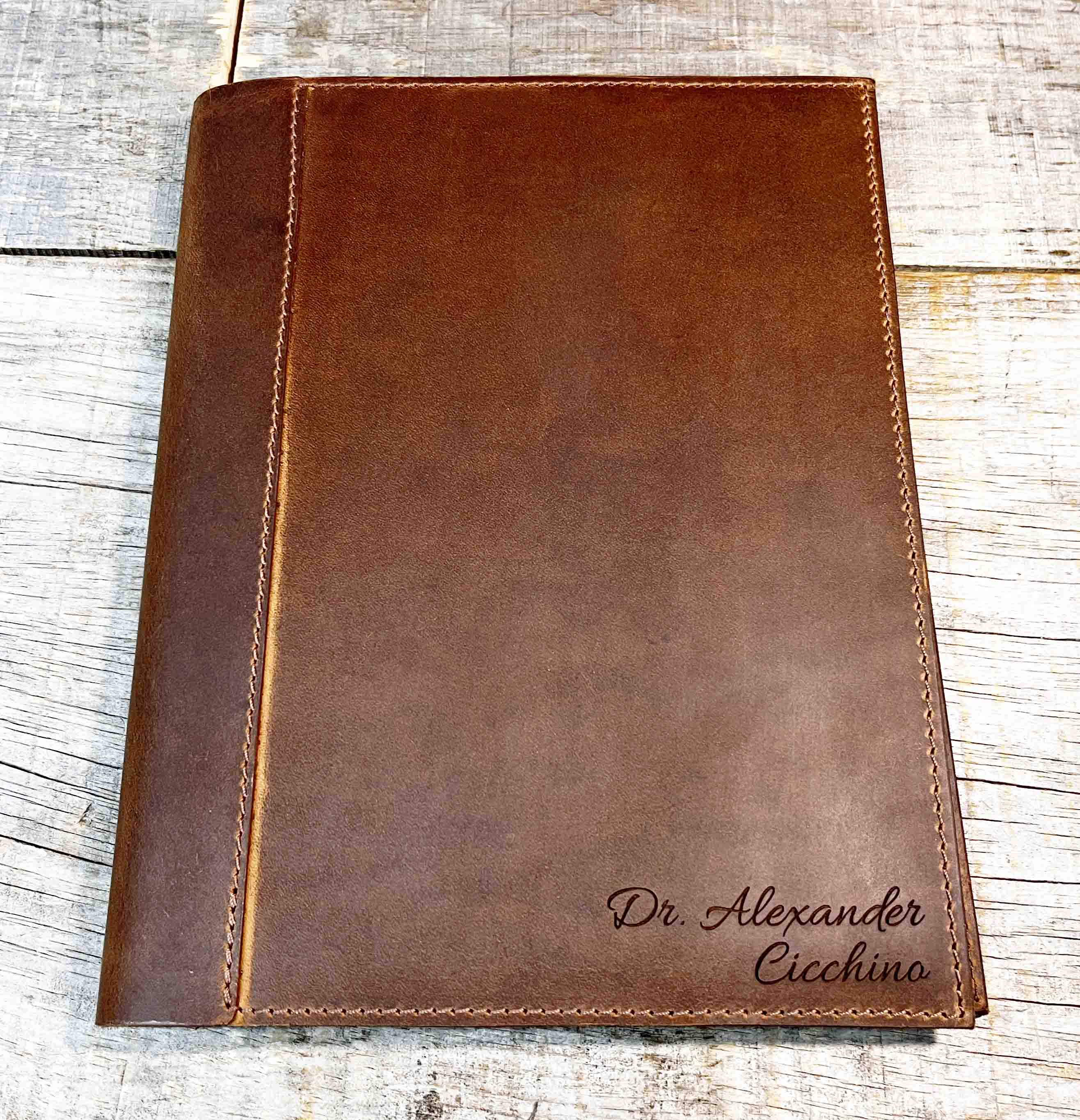 A brown, custom-engraved leather notebook cover featuring a geometric unicorn head design. The notebook is resting on an antique-style, ornate wooden desk alongside a glass inkwell and a feathered quill pen, creating a sophisticated and vintage aesthetic.