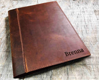 A brown, custom-engraved leather notebook cover featuring a geometric unicorn head design. The notebook is resting on an antique-style, ornate wooden desk alongside a glass inkwell and a feathered quill pen, creating a sophisticated and vintage aesthetic.